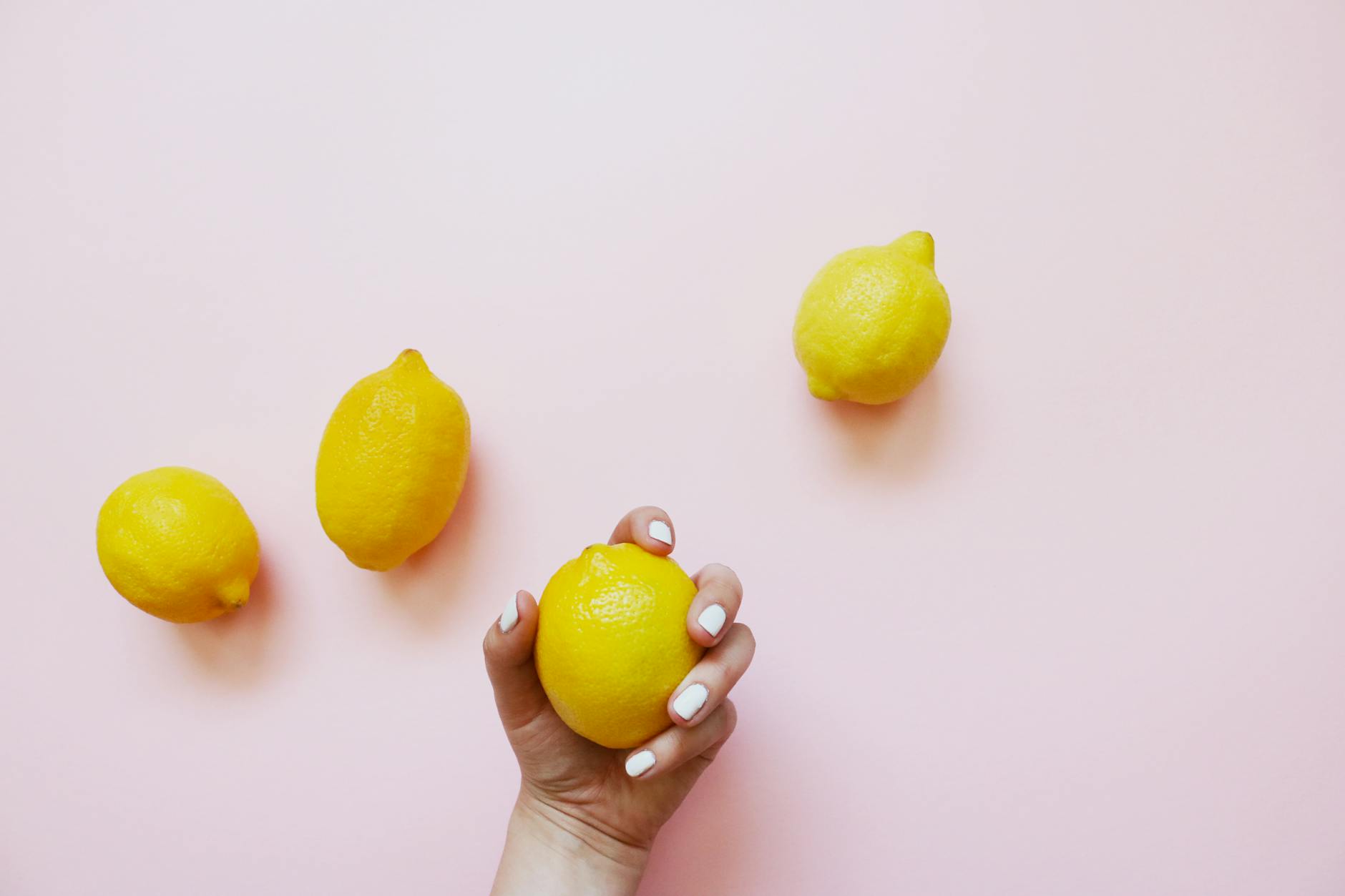 Eine Hand mit weißem Nagellack hält eine Zitrone auf zartem rosa Hintergrund