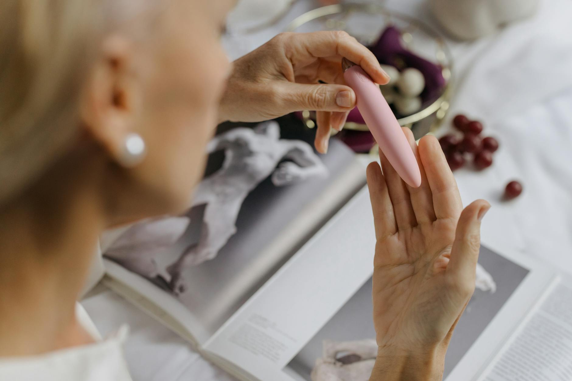 Nahaufnahme einer Frau, die einen rosa Vibrator hält, mit Kunstbüchern im Hintergrund, das persönliche und intime Themen betont.
