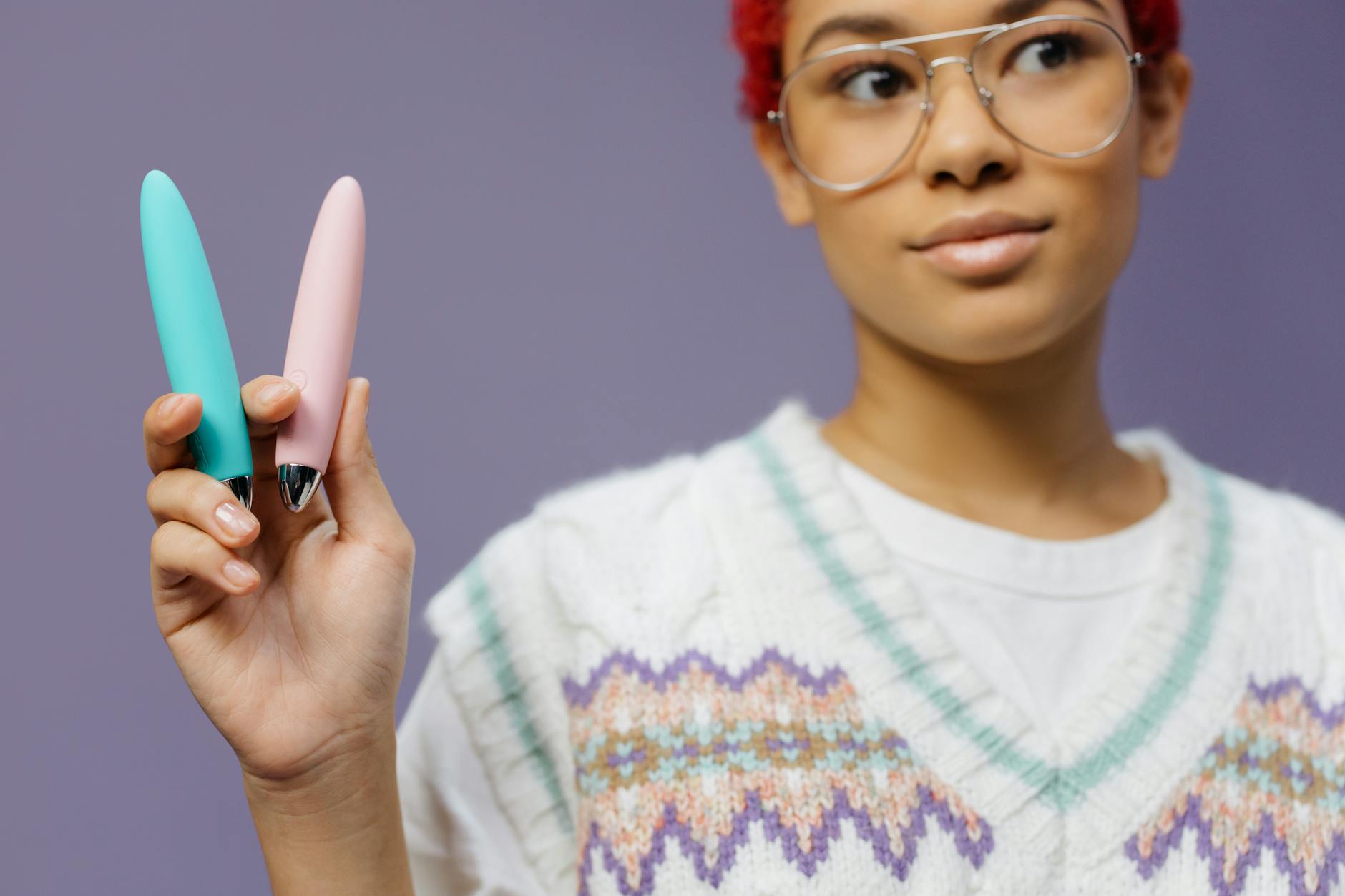 Eine Frau mit Brille hält zwei bunte Vibratoren vor einem lila Hintergrund.